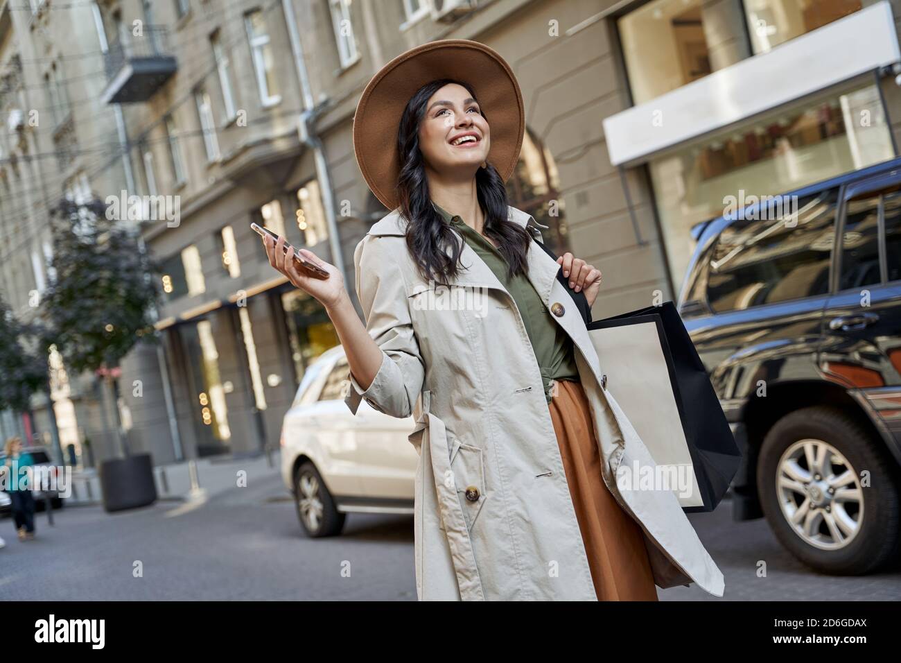 Zufrieden nach dem Einkaufen. Junge schöne stilvolle Frau in Hut mit Einkaufstasche hält ihr Smartphone, Blick zur Seite und lächelnd beim Gehen auf den Straßen der Stadt. Mode, Lifestyle Stockfoto