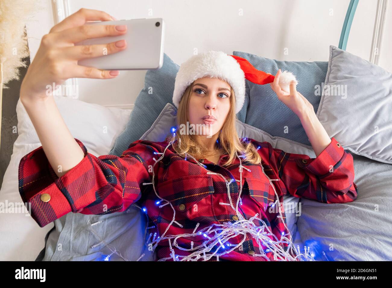 Aufgeregt junge Frau in santa Hut in einer Girlande gewickelt Selfie-Foto auf dem Handy, das auf dem Bett liegt Stockfoto