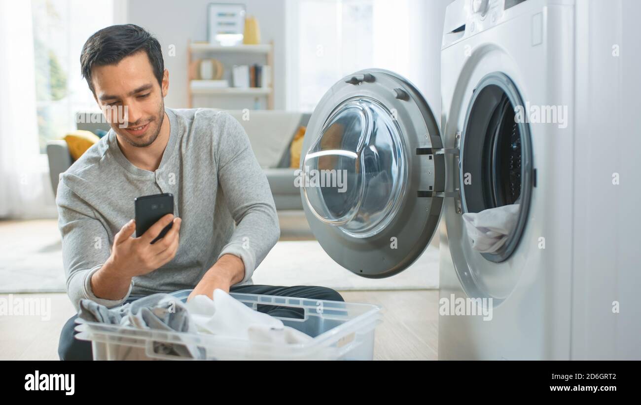 Schöner lächelnder junger Mann in grauer Jeans und Mantel sitzt vor einer  Waschmaschine und nutzt sein Smartphone. Er lädt Waschmaschine mit  schmutziger Wäsche Stockfotografie - Alamy