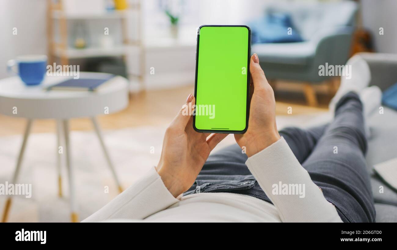 Junge Frau zu Hause Laying auf einer Couch mit Green Mock-up-Bildschirm Smartphone. Mädchen mit Chroma Key Handy, Internet-Browsing, Posting on Stockfoto