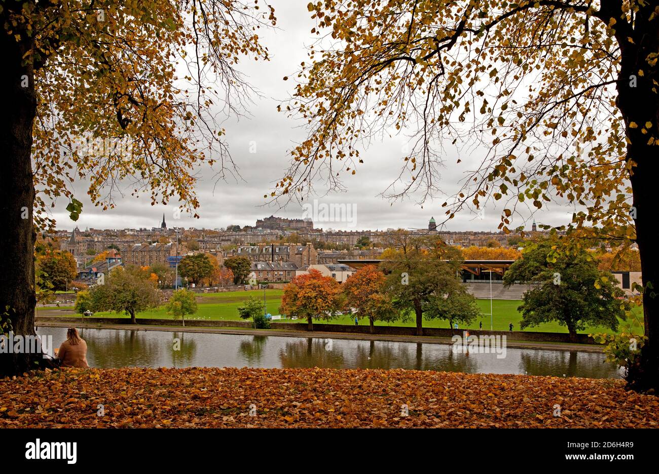 Inverleith Park, Edinburgh, Schottland, Großbritannien. 17. Oktober 2020. An einem bewölkten Tag verlieren die Bäume ihre Herbstfarben in Inverleith mit der Skyline der Innenstadt im Hintergrund.eine Dame sitzt unter einem Baum und bewundert den Blick über den Teich. Quelle: Arch White/Alamy Live News Stockfoto