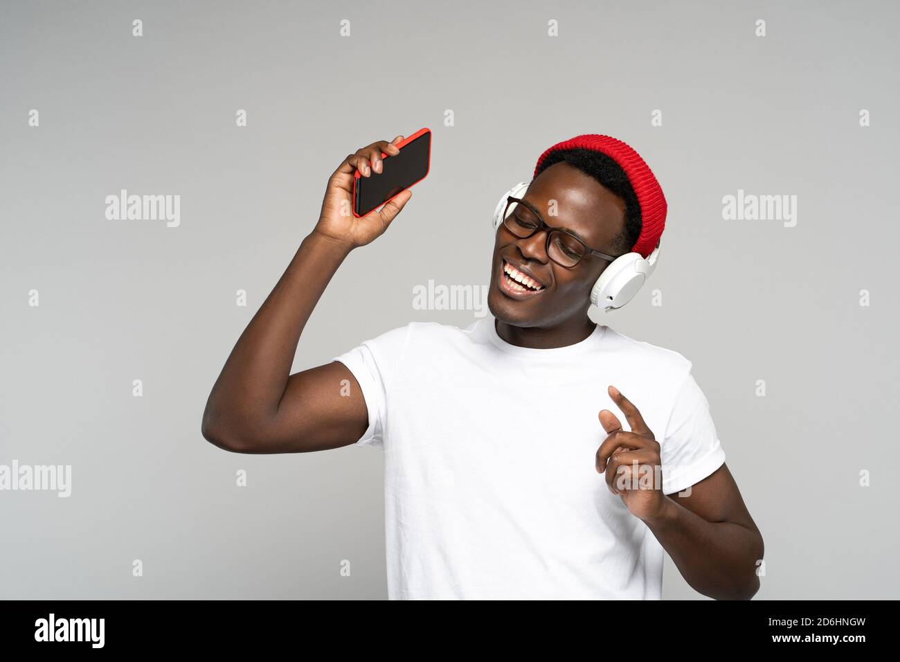 Afroamerikanischer Mann mit Kopfhörern tragen roten Hut, genießen Sie das neue Album des Künstlers zu hören, tanzt, macht Bewegungen zur Musik, hält mobile Phon Stockfoto