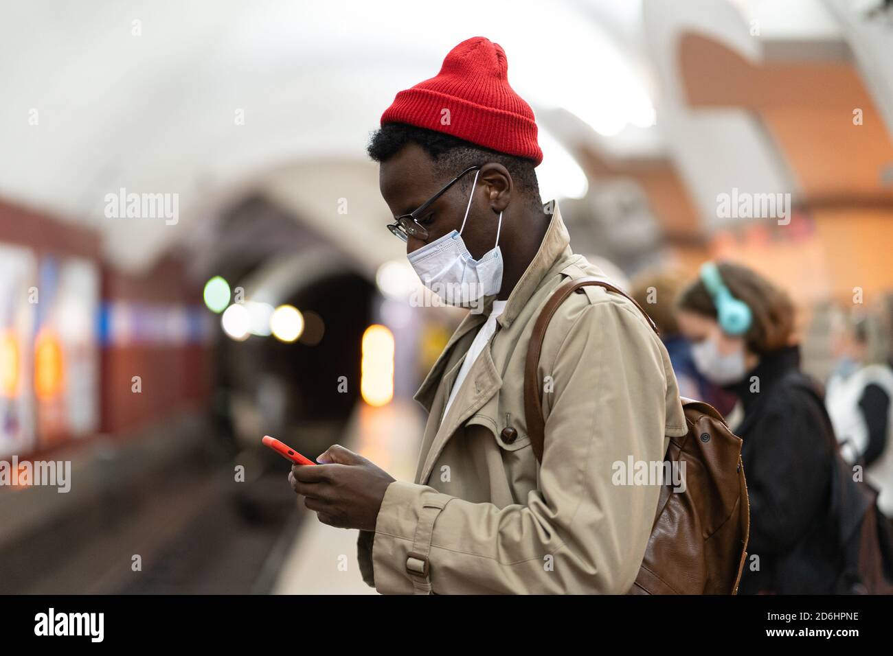 Schwarzer Tausendjähriger Mann im Trenchcoat, roter Hut mit Gesichtsmaske als Schutz gegen covid-19, Grippe-Virus, wartet auf den Zug an der U-Bahn-Station, usi Stockfoto