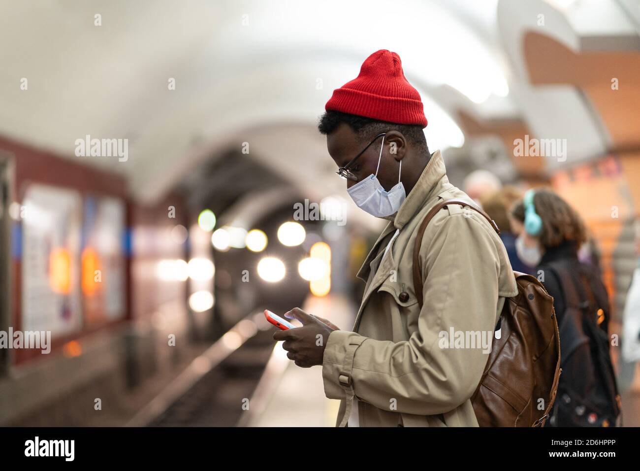 Schwarzer Tausendjähriger Mann im Trenchcoat, roter Hut mit Gesichtsmaske als Schutz gegen covid-19, Grippe-Virus, wartet auf den Zug an der U-Bahn-Station, usi Stockfoto