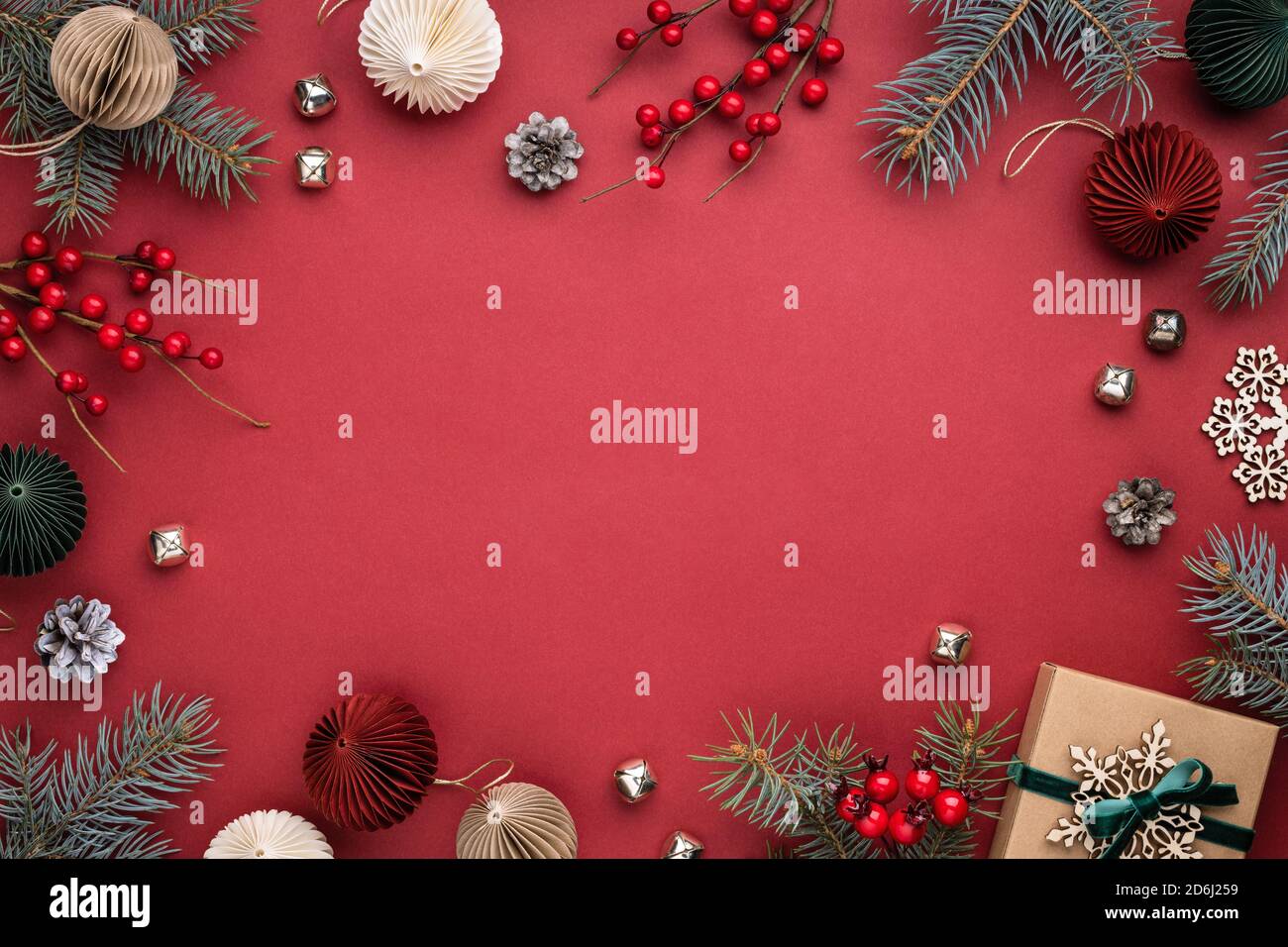 Weihnachtsrahmen mit Geschenkbox, Papierdekorationen, Fichtenzweigen und Beeren auf rotem Hintergrund. Feiertagsgrenze in Erdfarben. Stockfoto