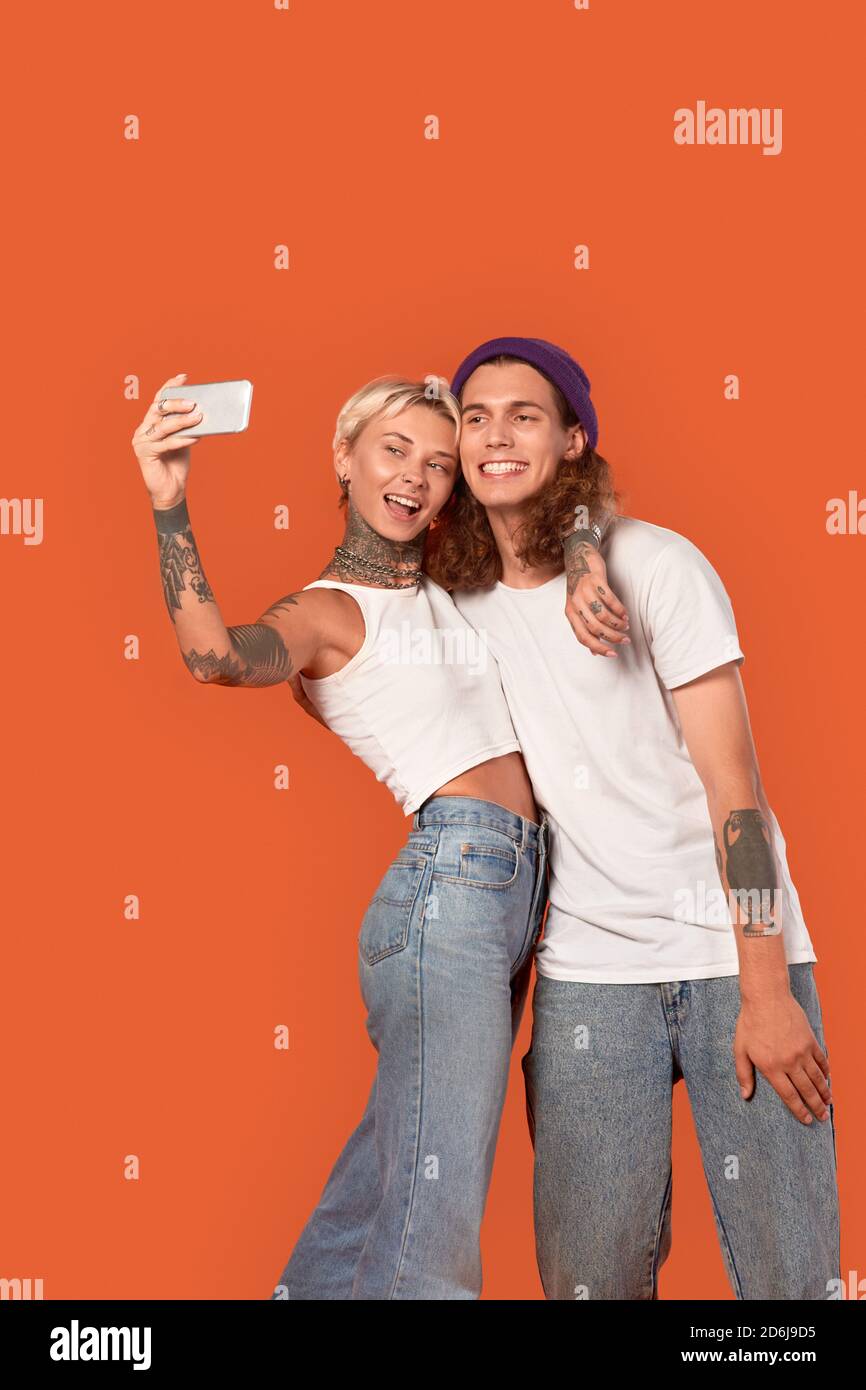 Freistil. Junge tätowierte Paar auf orange Umarmung isoliert stehend unter Selfie auf Smartphone posiert Kamera verspielt Stockfoto