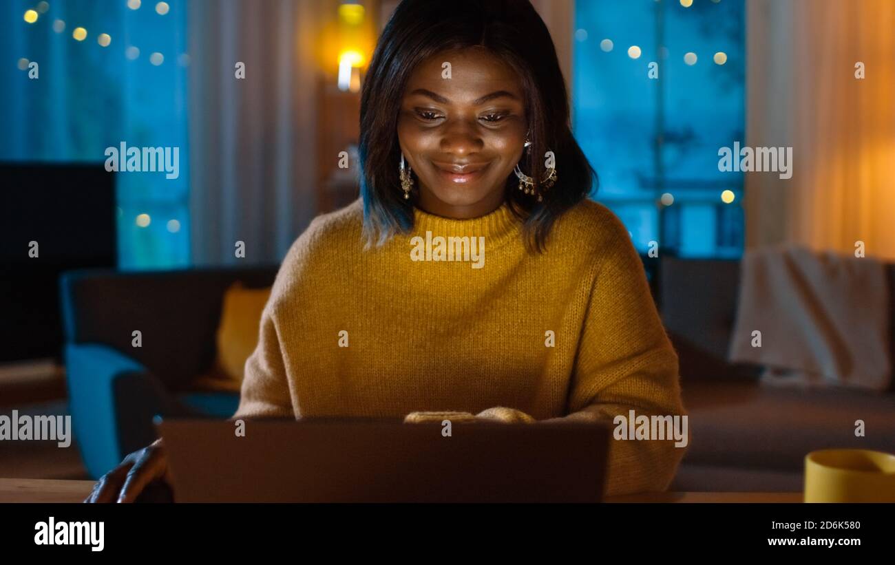 Portrait von schönen lächelnden schwarzen Mädchen arbeiten auf einem Laptop, während sie an ihrem Schreibtisch zu Hause sitzen. Am Abend arbeitet Creative Woman an einem Computer in Stockfoto