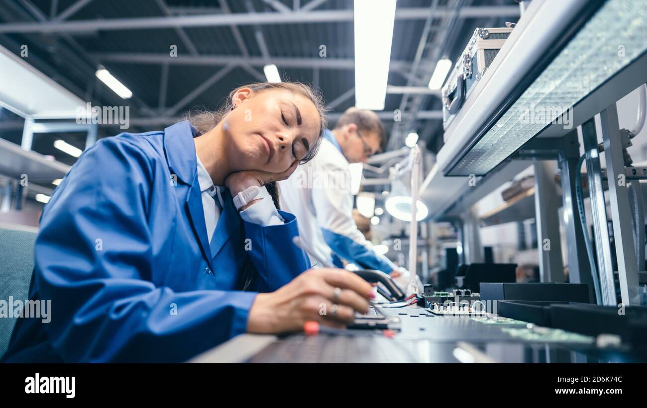 Aufnahme einer müden schlafenden Frau in blauem Arbeitsmantel an ihrem Arbeitsplatz in der Elektronikfabrik. High Tech Factory Facility mit mehr Mitarbeitern in der Stockfoto