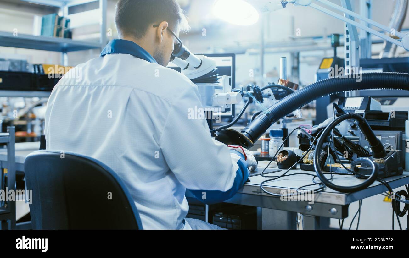 Rückansicht Aufnahme eines Arbeiters in der Elektronikfabrik in weißem Arbeitsmantel inspiziert eine Leiterplatte durch ein digitales Mikroskop. High-Tech-Fabrik Stockfoto