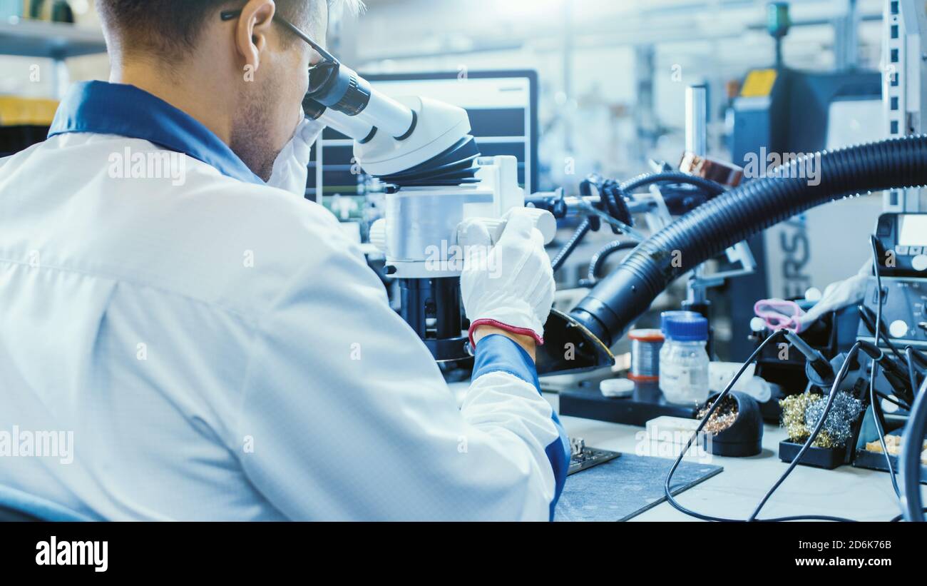 Arbeiter in der Elektronikfabrik in White Work Coat löten eine Leiterplatte über ein digitales Mikroskop. High-Tech-Fabrik. Stockfoto