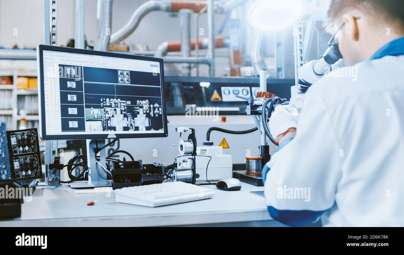 Arbeiter in der Elektronikfabrik in weißer Arbeitsschicht inspiziert eine Leiterplatte auf einem Computerbildschirm, der mit einem digitalen Mikroskop verbunden ist. Hoch Stockfoto