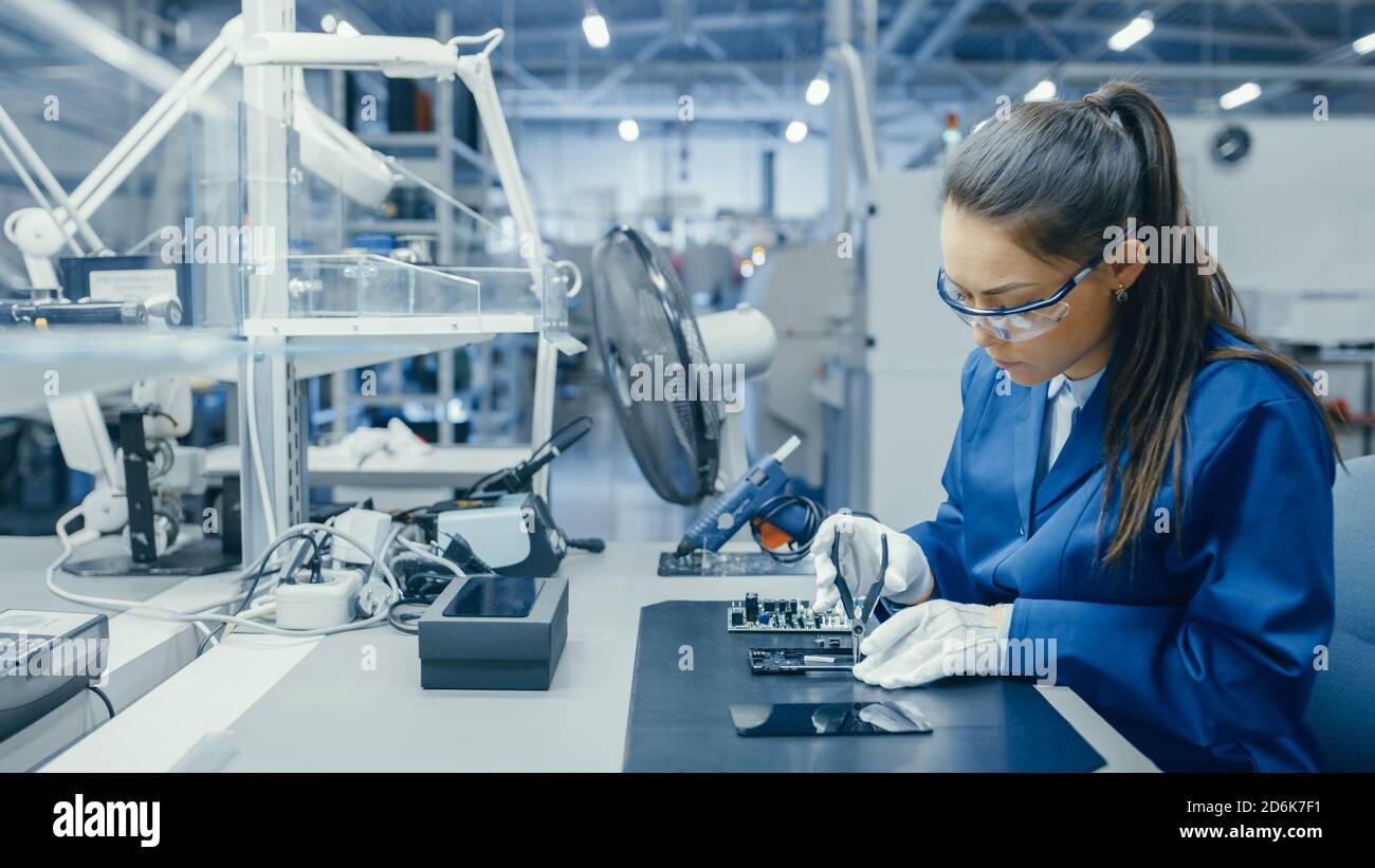Young Female Blue and White Work Coat verwendet Zange, um gedruckte Leiterplatte für Smartphone zu montieren. Arbeiter in der Elektronikfabrik in einer High-Tech Stockfoto