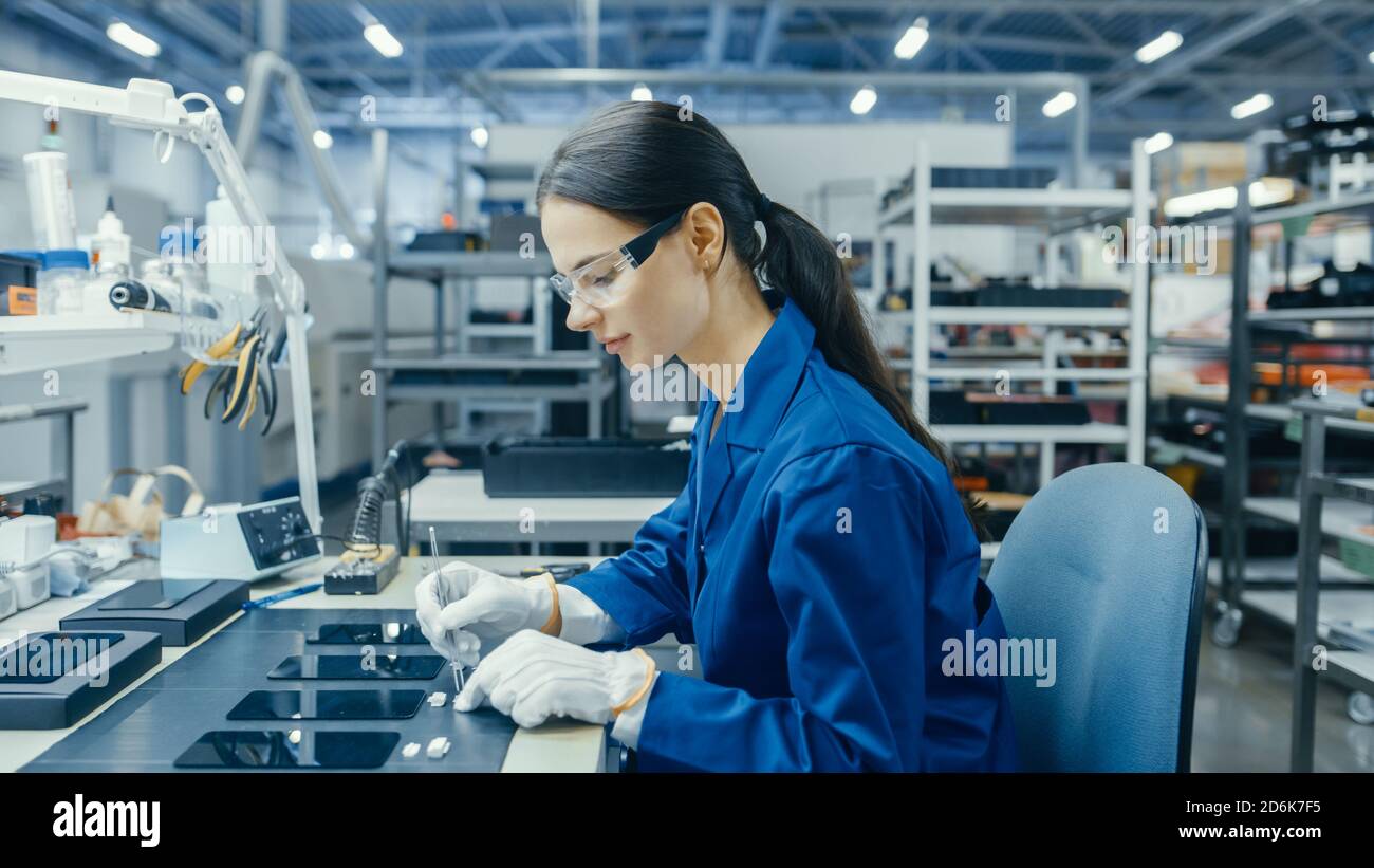 Young Female in Blue Work Coat ist die Montage von Leiterplatten für Smartphones. Arbeiter in der Elektronikfabrik in einer High-Tech-Fabrik. Stockfoto