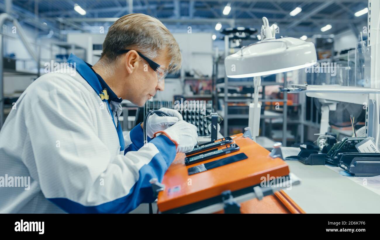 Man in Blue Work Coats verwendet eine Pinzette, um Leiterplatten für Smartphones zu montieren. Arbeiter in der Elektronikfabrik in einer High-Tech-Fabrik Stockfoto