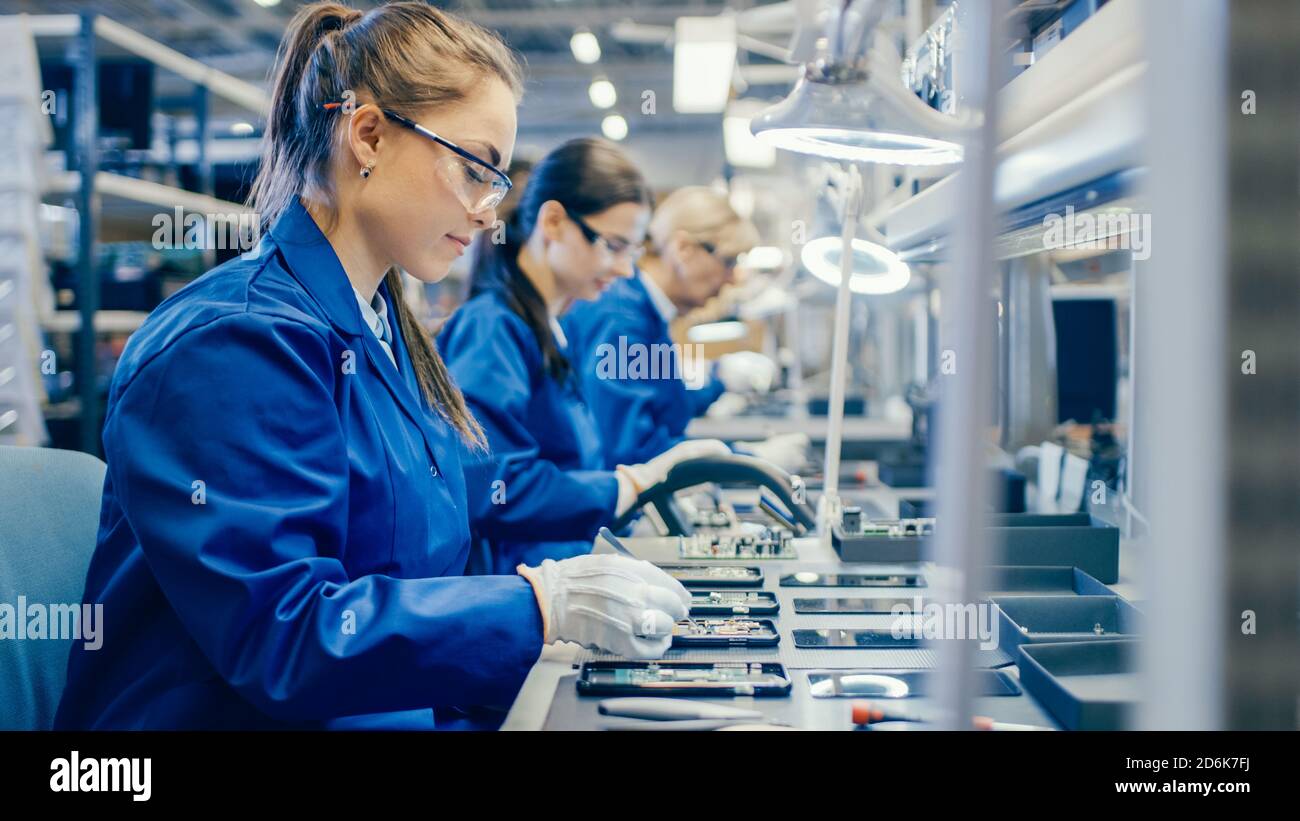 Arbeiterinnen in der Elektronikfabrik Blue Work Coat und Schutzbrille Montage von Leiterplatten für Smartphones mit Pinzette. Hightech Stockfoto