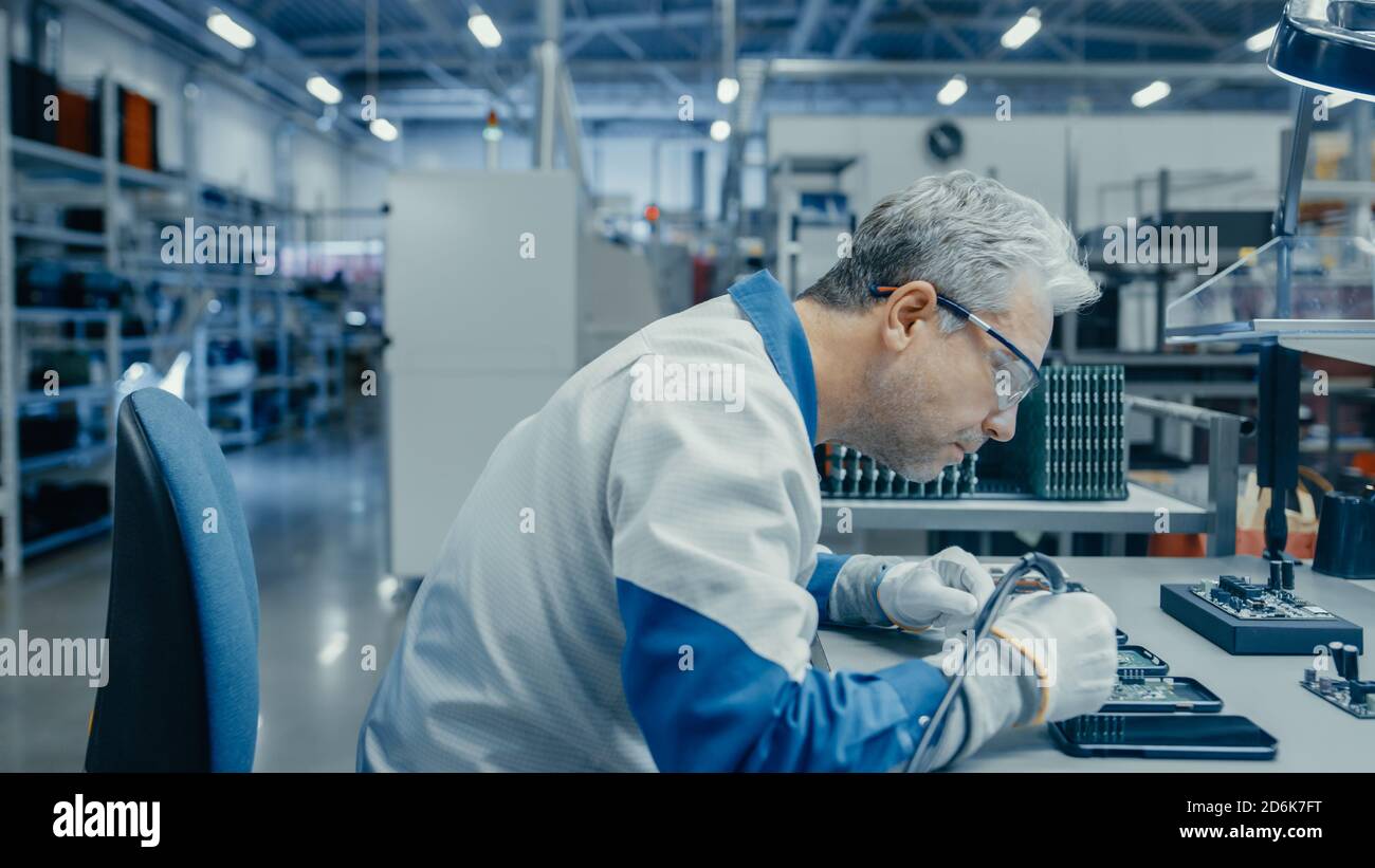 Senior man in Blau - White Work Coat verwendet Zange, um gedruckte Leiterplatte für Smartphone zu montieren. Arbeiter in der Elektronikfabrik in einer High-Tech Stockfoto
