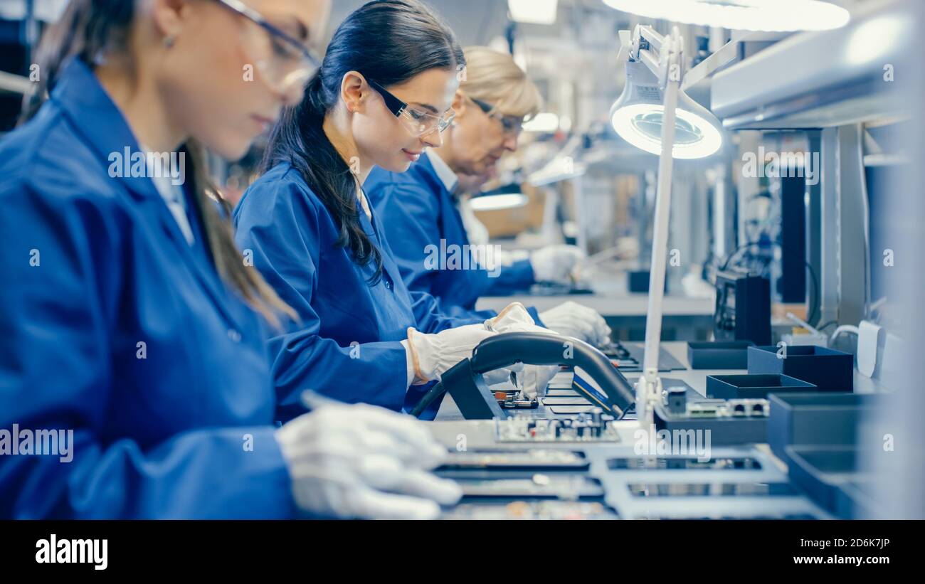 Arbeiterinnen in der Elektronikfabrik Blue Work Coat und Schutzbrille Montage von Leiterplatten für Smartphones mit Pinzette. Hightech Stockfoto