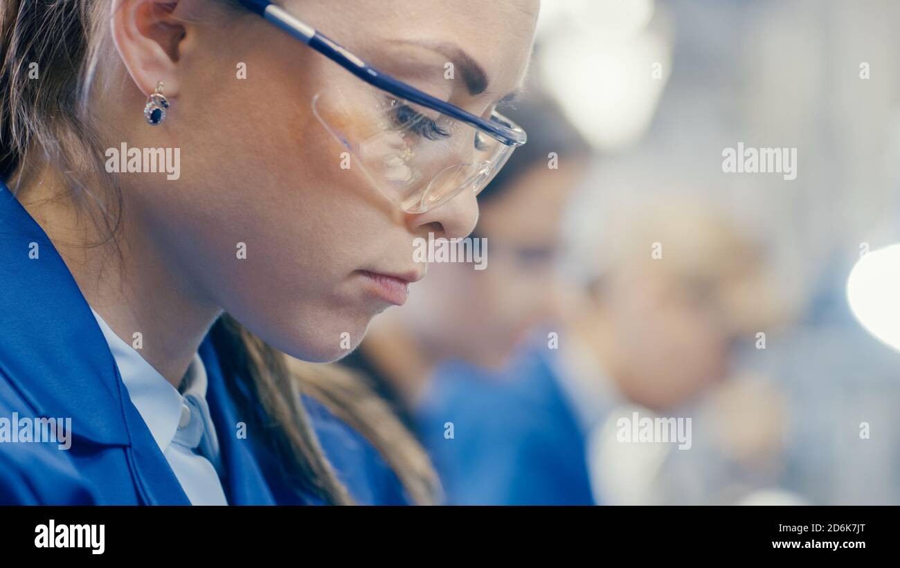 Nahaufnahme einer Arbeiterin in der Elektronikfabrik in blauer Arbeitskleidung und Schutzbrille schaut nach unten und arbeitet. High-Tech-Fabrik Stockfoto