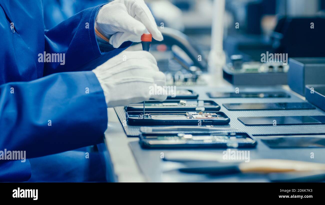 Nahaufnahme einer Arbeiterin in der Elektronikfabrik in blauem Arbeitsmantel und Schutzbrille Montage von Smartphones mit Schraubendreher. High-Tech-Fabrik Stockfoto