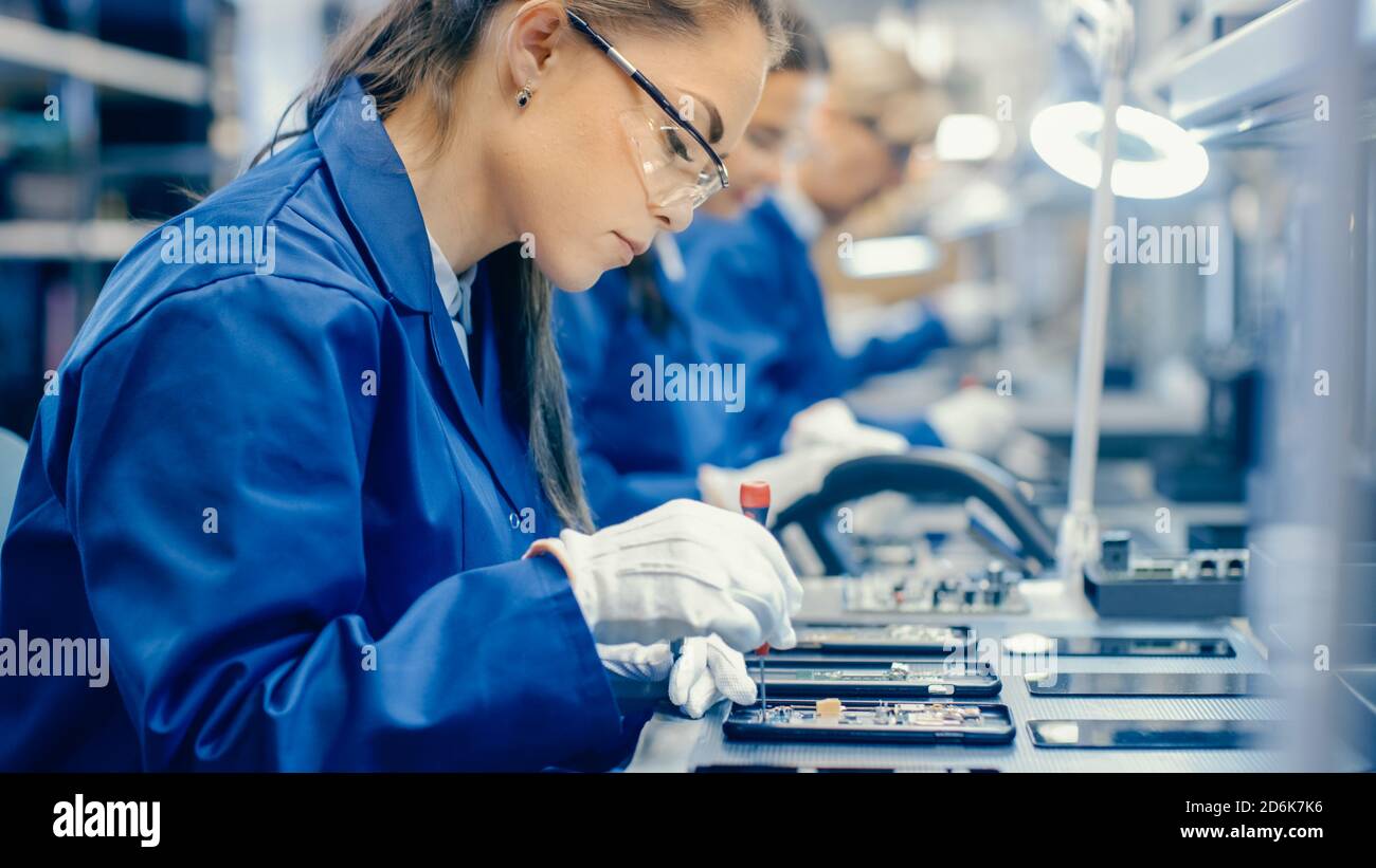 Arbeiterinnen in der Elektronikfabrik Blue Work Coat und Schutzbrille Montage von Leiterplatten für Smartphones mit Pinzette. Hightech Stockfoto