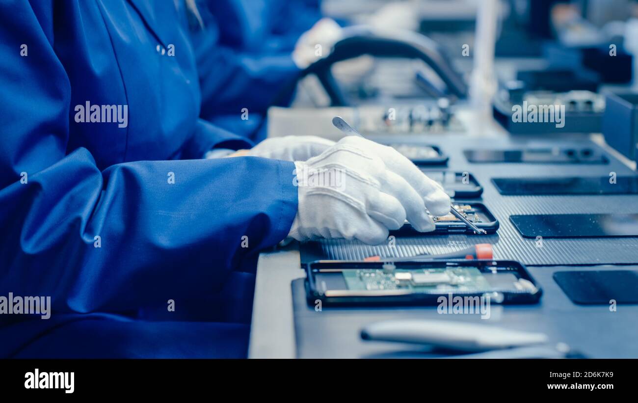 Nahaufnahme einer Arbeiterin in der Elektronikfabrik in blauem Arbeitsmantel und Schutzbrille Montage von Smartphones mit Schraubendreher. High-Tech-Fabrik Stockfoto