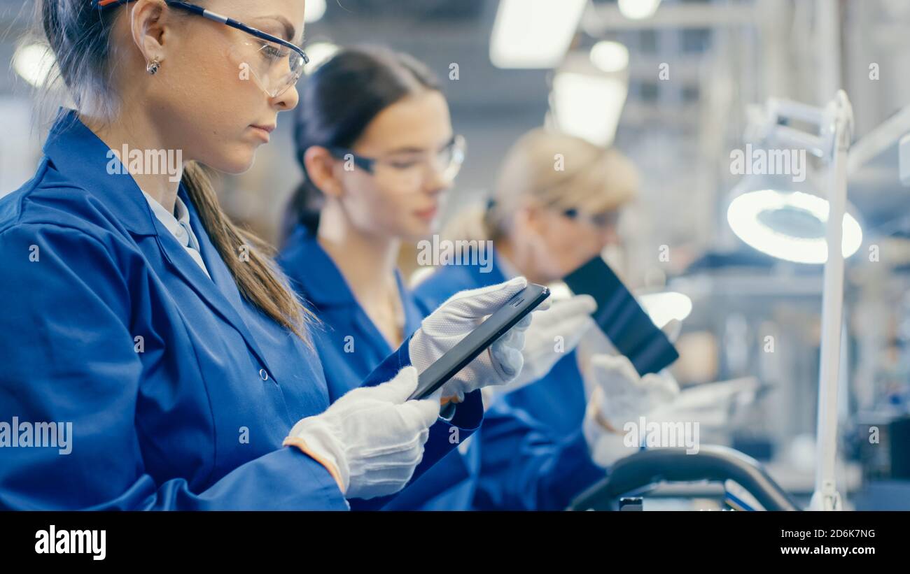 Woman Electronics Factory Worker in Blue Work Coat und Schutzbrille nimmt ein Smartphone-Bildschirm und führt eine Qualitätsprüfung. High-Tech-Fabrik Stockfoto