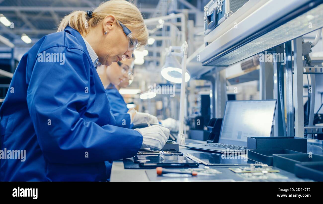 Weibliche Elektronik-Fabrik Arbeiter in Blue Work Coat und Schutzbrille ist Montage Smartphones mit Schraubendreher. High-Tech-Fabrik mit Stockfoto