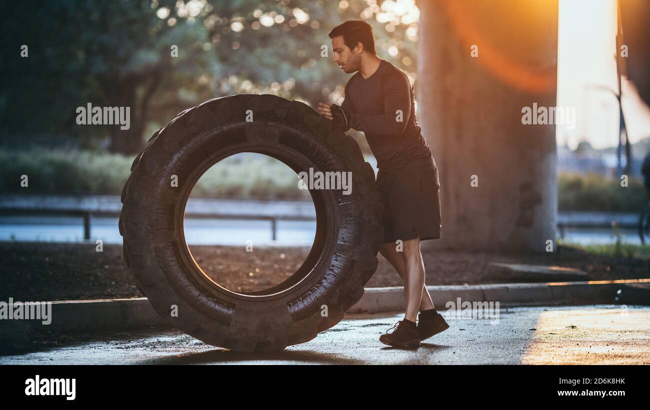 Starker muskulöser Fit junger Mann macht Übungen auf der Straße. Er hebt und rollte einen großen schweren Reifen in einer städtischen Umgebung. Stockfoto