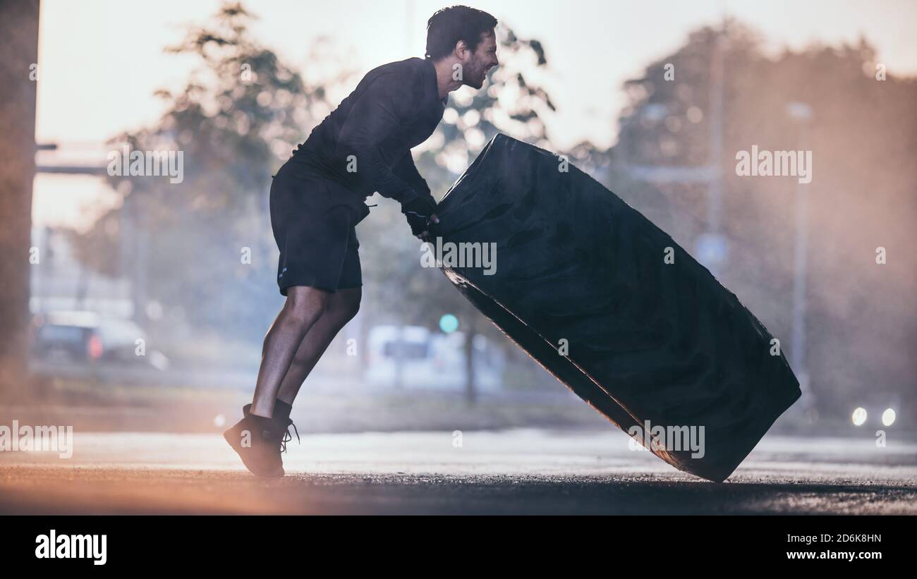 Starker muskulöser Fit junger Mann macht Übungen auf der Straße. Er dreht einen großen schweren Reifen in einer städtischen Umgebung. Stockfoto