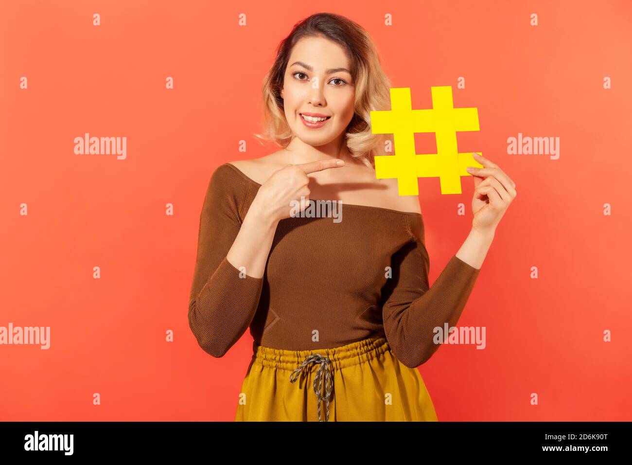 Positive attraktive Frau blogger zeigt Finger auf große gelbe Hashtag in der Hand, Tagging Beiträge in sozialen Netzwerken, Popularität. Innenaufnahme im Studio Stockfoto