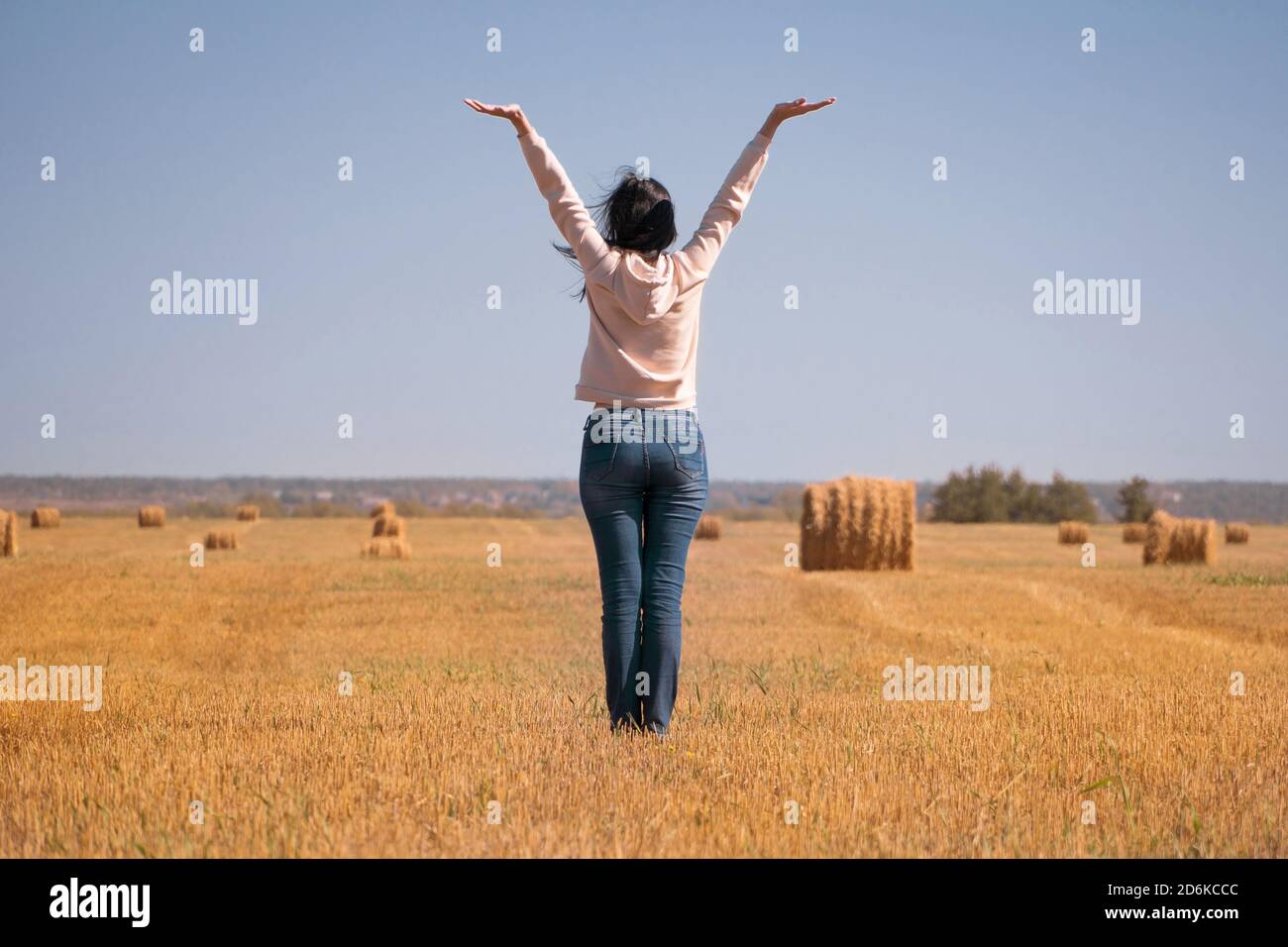 Ein glückliches, freies, unabhängiges, verträumtes Mädchen auf dem Feld hob ihre Hände in den Himmel. Eine Frau in einer Jacke und Jeans bei der Ernte Rückansicht. Silhouette o Stockfoto