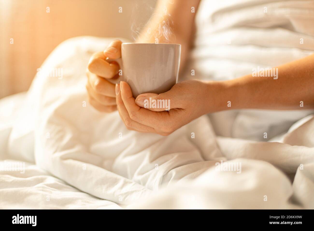 Frau Hand hält Tasse Kaffee oder Tee im Bett auf weiße Decke. Kaffee am Morgen Konzept Stockfoto