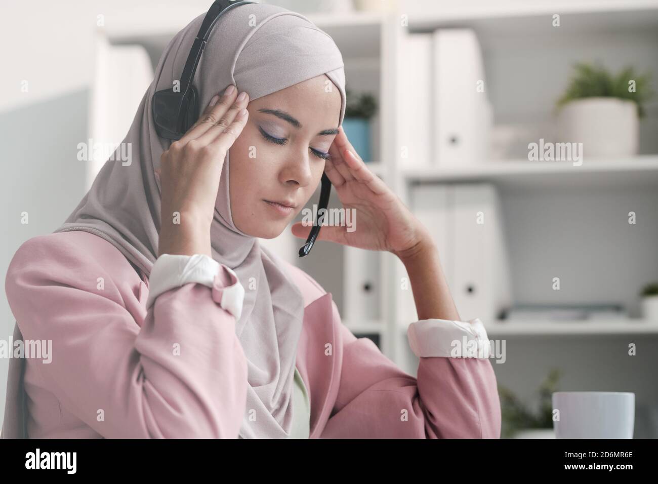 Junge müde muslimische Geschäftsfrau in Hijab und Headset berühren sie Tempel Stockfoto