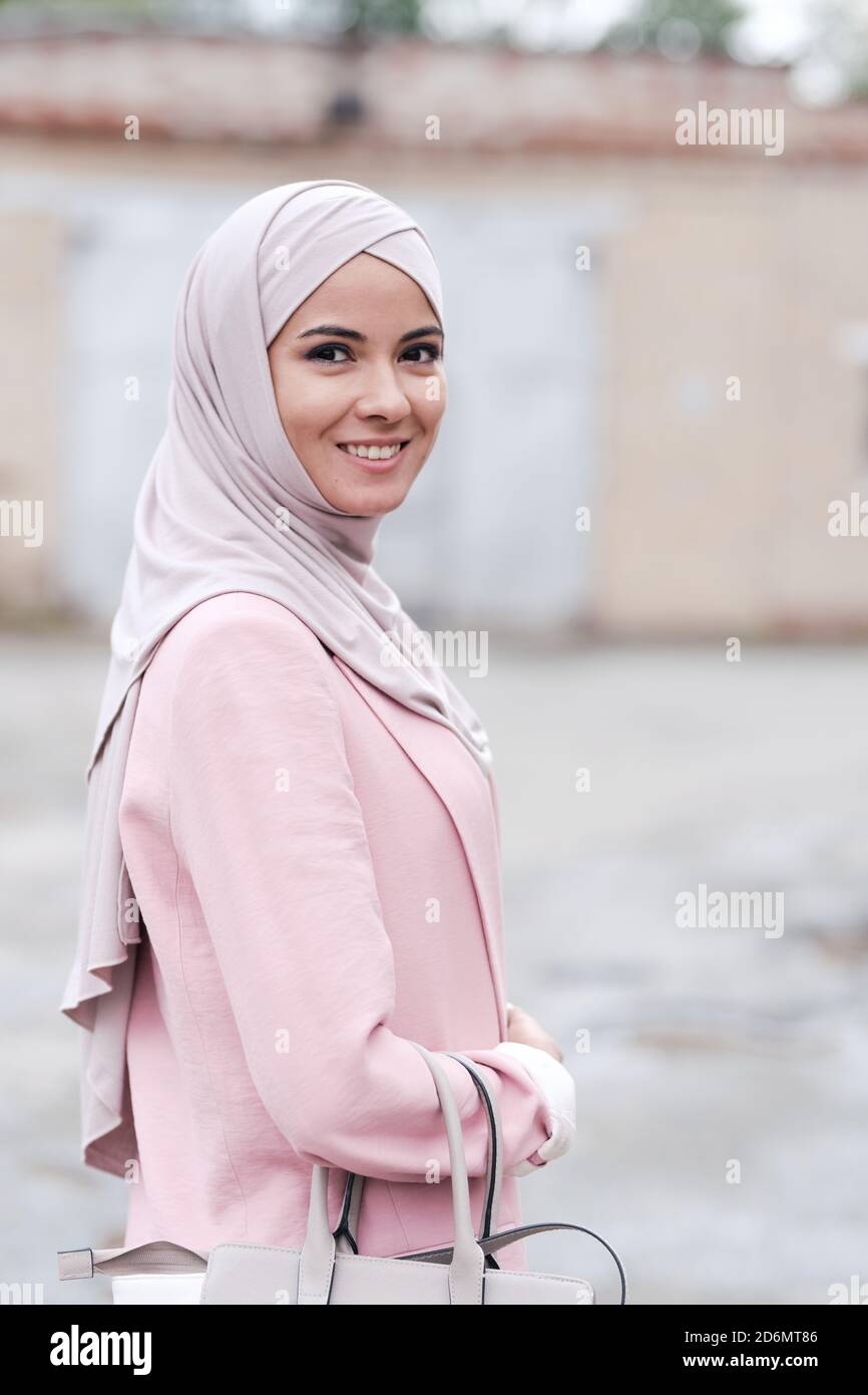 Junge fröhliche muslimische Frau in Hijab und rosa Strickjacke halten Handtasche Stockfoto