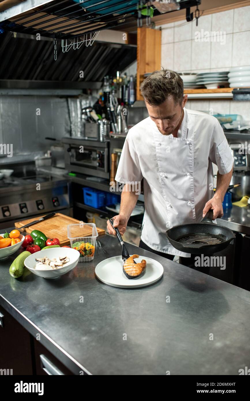 Junger männlicher Koch in weißer Uniform Putting Stück gebraten Lachs auf dem Teller Stockfoto