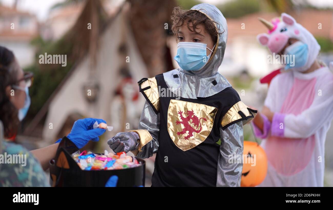 Kinder tragen Kostüme und Gesichtsmasken aus Trick oder Behandlung an Halloween 2020. Eine Person gibt Süßigkeiten mit Handschuhen aus. Stockfoto