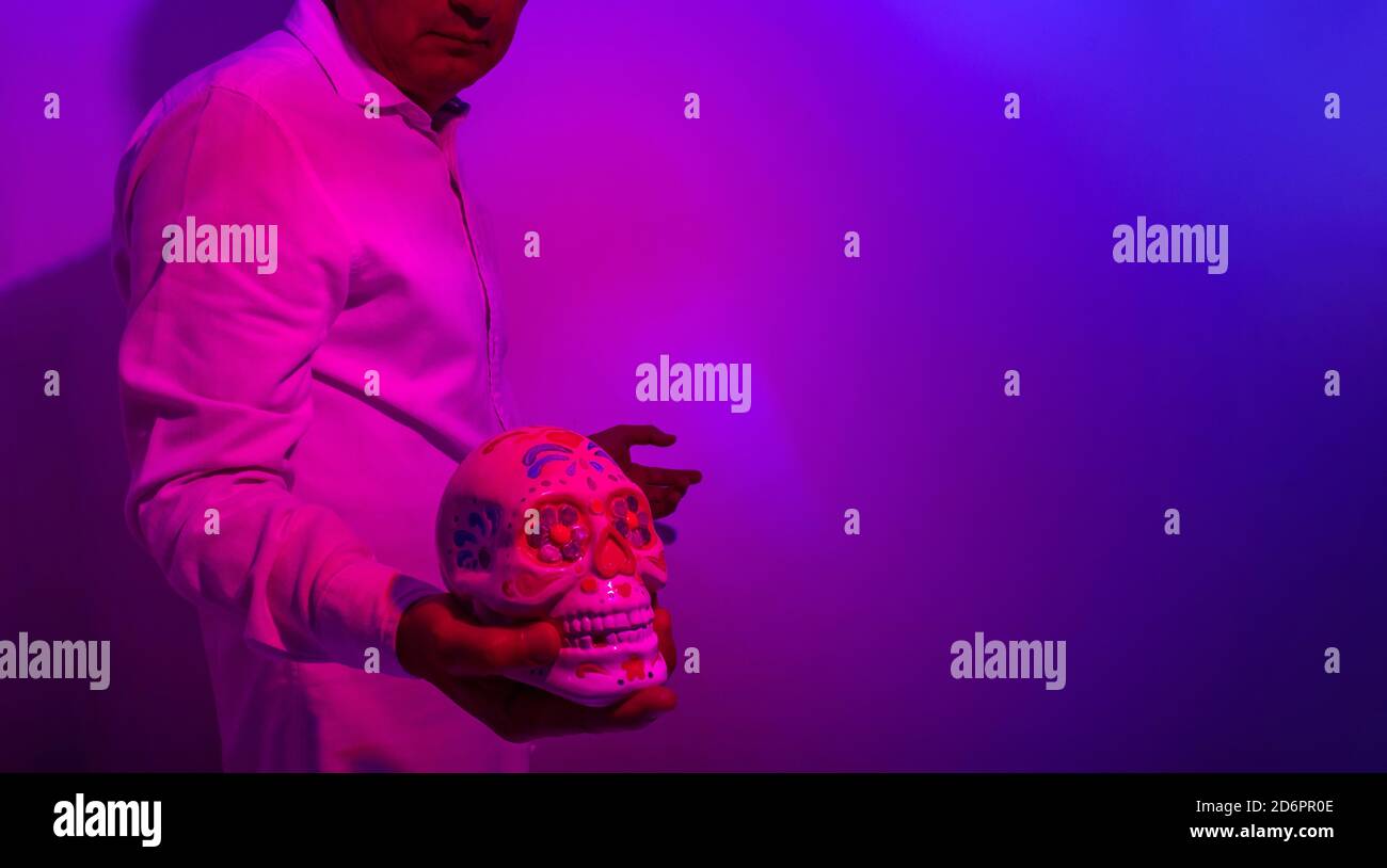 Ein Mann, dessen Gesicht nicht erkennbar ist, mit einem bemalten Totenschädel aus dem Tag der Toten oder halloween in der Hand. Fotografieren Sie in violetten Tönen. Selektiver Fokus Stockfoto
