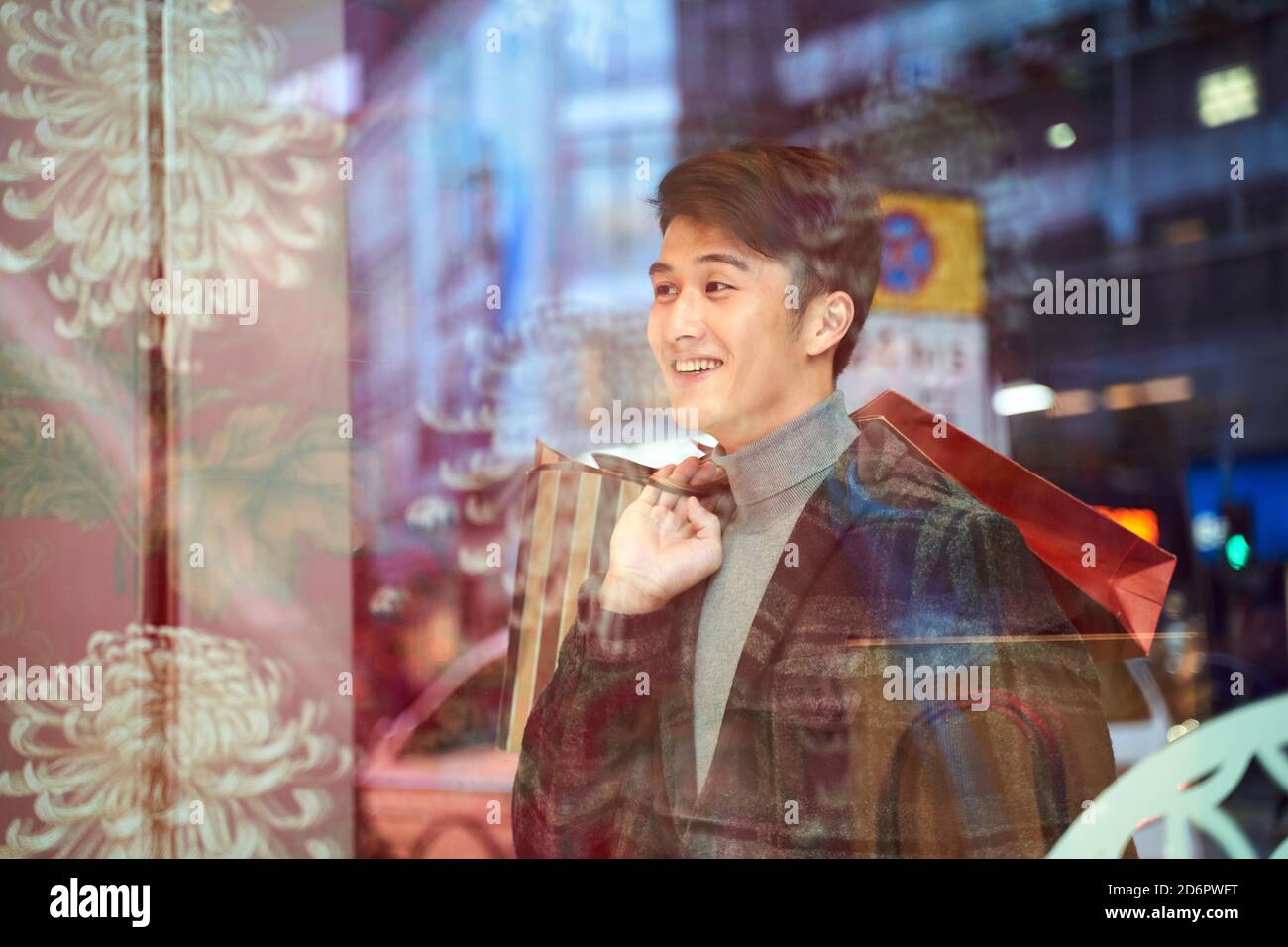 Glücklicher junger asiatischer Mann, der in das Schaufenster schaut und lächelt Moderne Stadt Stockfoto