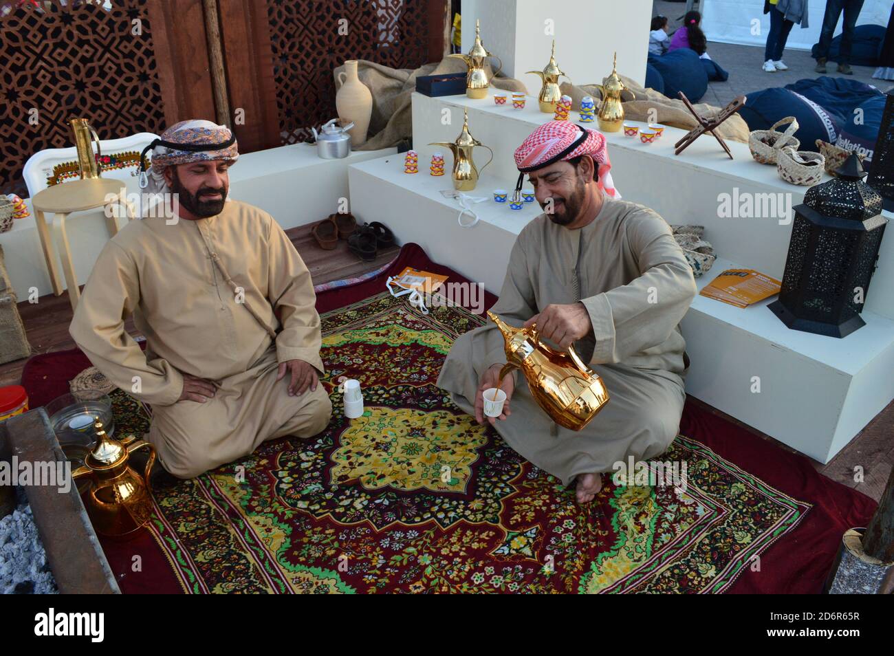 Abu Dhabi, VAE - 2019. Zwei arabische Männer, die traditionell angezogen waren, tranken Kaffee aus einer glänzenden Kaffeekocher und saßen auf dem farbenfrohen Teppich Stockfoto