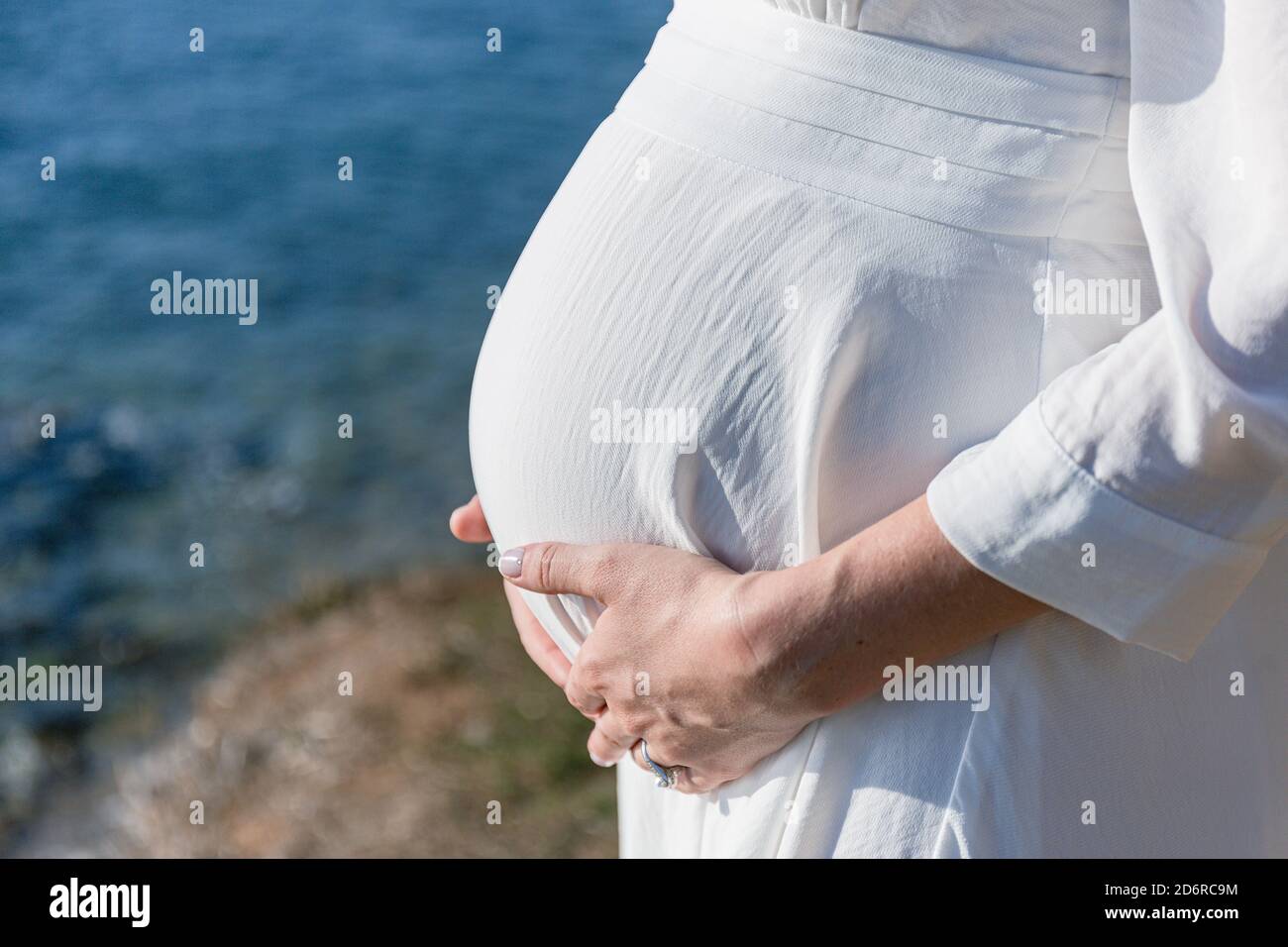 Eine schwangere Frau in einem weißen Kleid hält ihre Hände auf ihrem Bauch. Das Konzept der Schwangerschaft, Mutterschaft, Vorbereitung und Erwartung. Nahaufnahme Stockfoto