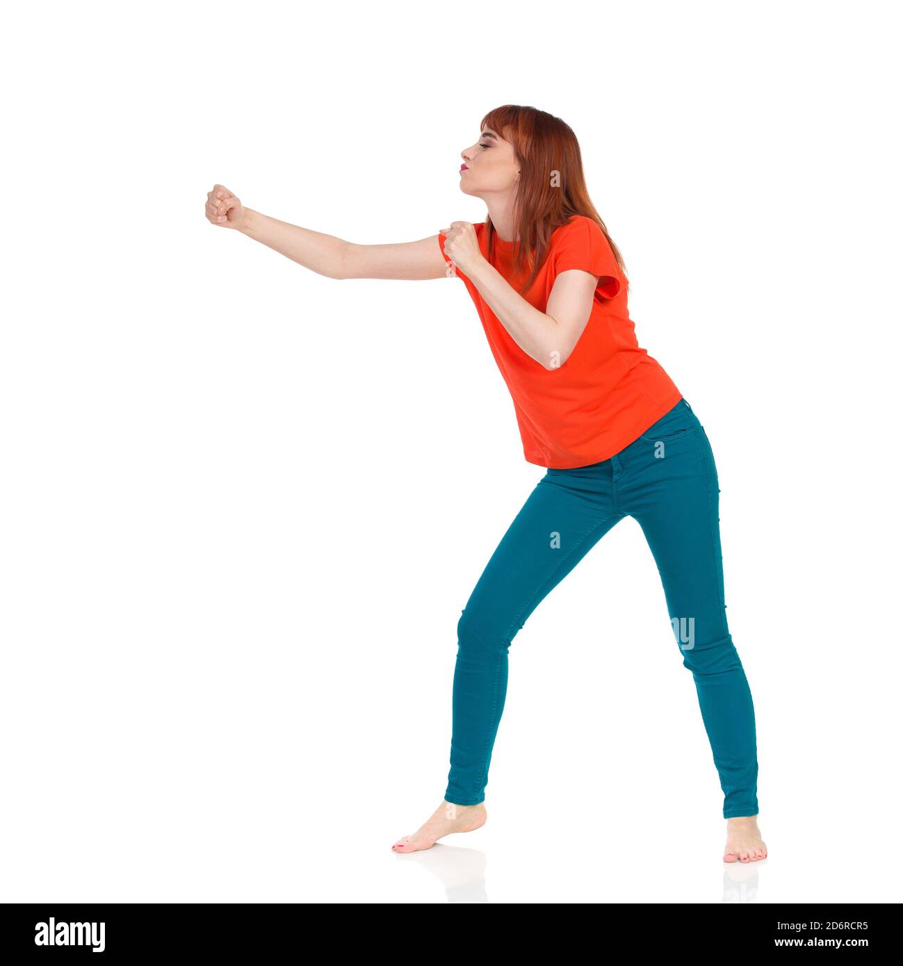 Junge Frau im orangefarbenen T-Shirt und grünen Jeans steht barfuß, klatscht ihre Faust und schaut weg. Studioaufnahme in voller Länge, isoliert auf Weiß. Stockfoto