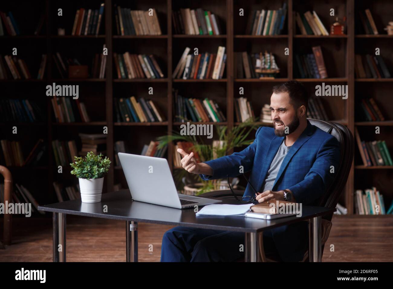 Wütend verrückt verärgerten Geschäftsmann in der Bibliothek Büro zeigt den Mittelfinger auf dem Laptop-Bildschirm. Anstrengende Verhandlungen durch Videokonferenz Stockfoto