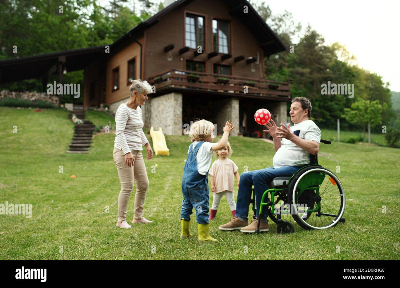 Kleine Kinder mit älteren Großeltern im Rollstuhl spielen mit einem Ball im Garten. Stockfoto