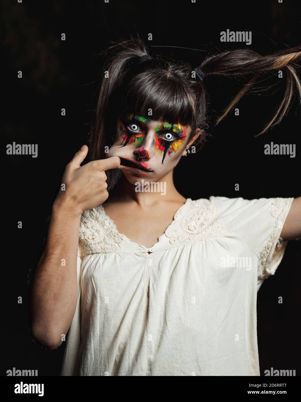 Gruselige junge Frau in bunten Kontaktlinsen und mit bemalt Gesicht stehen in dunklen Wald und lächeln gruselig beim Schauen An Halloween vor der Kamera Stockfoto