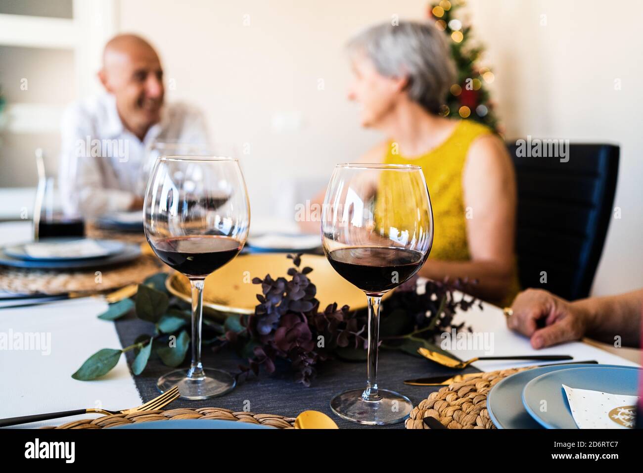 Esstisch serviert mit Gläsern und Tellern gegen verschwommenes Leuchten Weihnachtsbaum am Heiligabend zu Hause Stockfoto