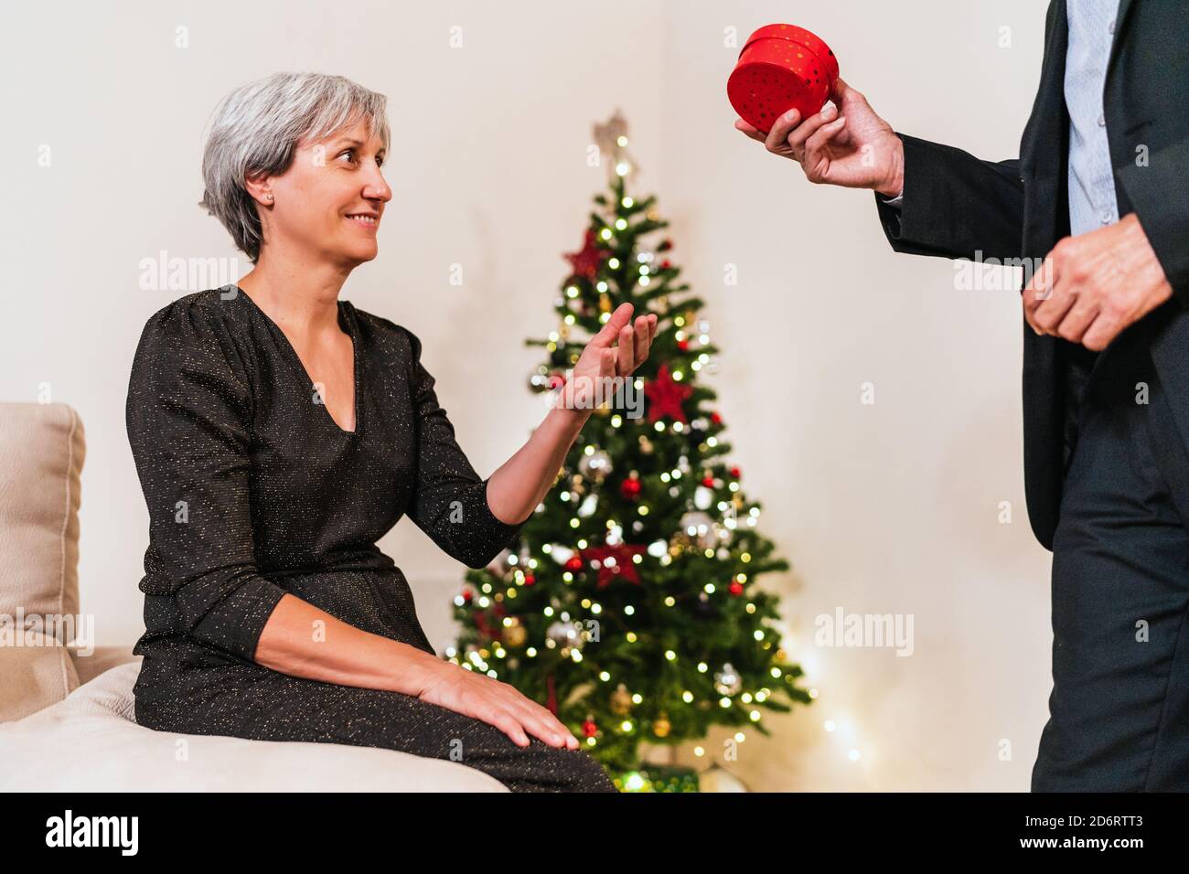 Seitenansicht des beschnittenen unkenntlichen Mannes, der während der Party zu Hause in einer Schachtel für eine liebende Frau auf dem Hintergrund des Weihnachtsbaums präsent ist Stockfoto