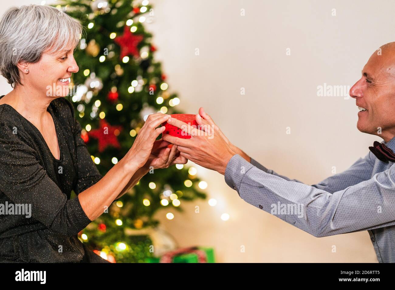Seitenansicht der fröhlichen reifen Mann geben Geschenk in der Box für liebevolle Frau auf dem Hintergrund von Weihnachtsbaum während der Party zu Hause Stockfoto