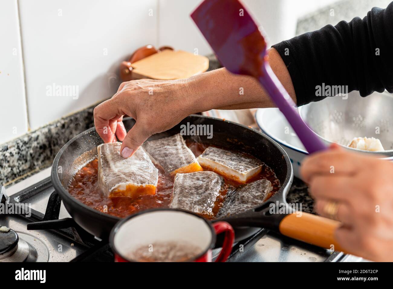 Crop anonyme Frau, die Kabeljau-Steaks in eine Pfanne mit kochender Sauce legt, während sie zu Hause ein typisch katalanisches Gericht Bacalao a la Catalana zubereitet Stockfoto