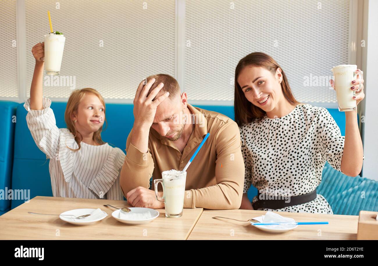 Glückliche Dreierfamilie hat Spaß im lokalen Café, Mutter und Tochter hoben ihre Gläser mit Cocktails, Vater sitzt zwischen ihnen und ruht seinen Kopf in der Hand Stockfoto