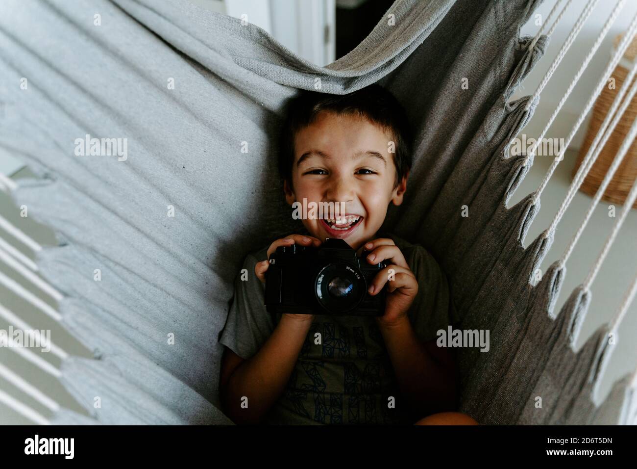 Von oben lachenden Jungen halten moderne professionelle Fotokamera während Ruhen auf hängende Hängematte in der Wohnung und Blick auf die Kamera Glücklich Stockfoto